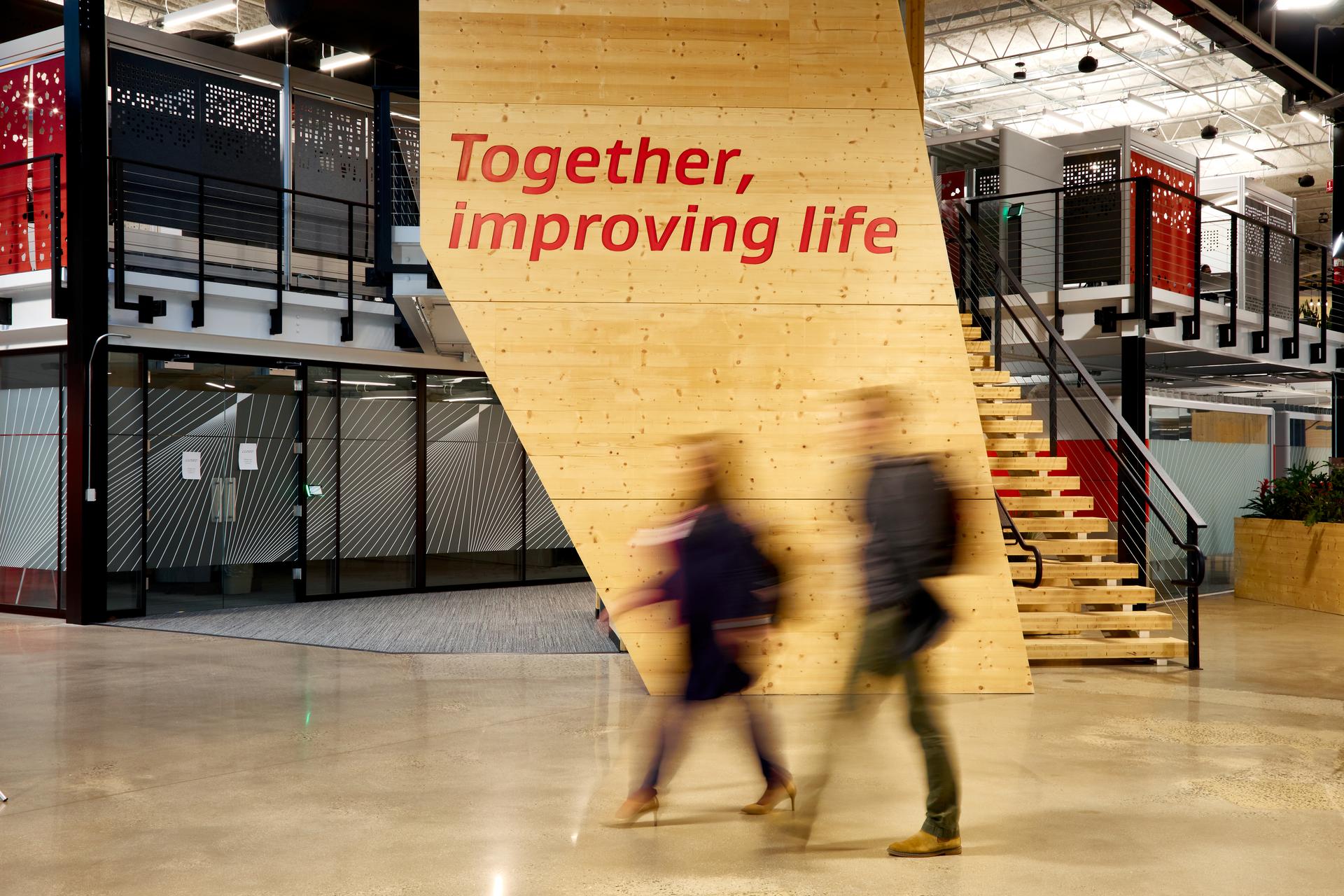 壁にTogether, improving lifeというブランドプロミスが掲げられたオフィスを歩くアソシエートたちのぼやけた姿