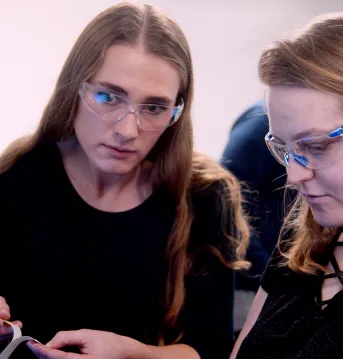 Two Associates wearing safety glasses closely examine a strip of material while working together in a lab